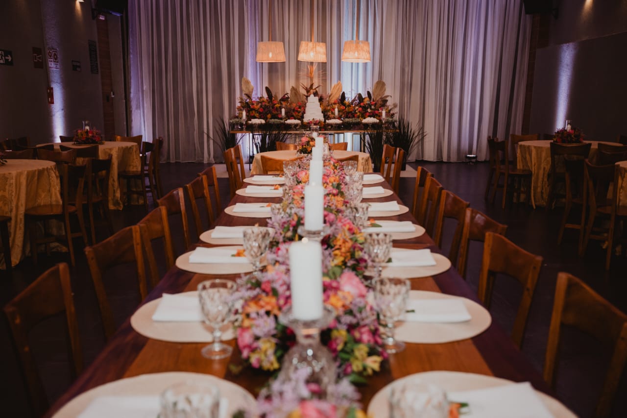 Mesa longa de madeira com flores rosas, taças de cristal e telão com logo do Jardins Caiçara